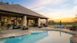 A pool with a stone patio, outdoor seating, and a building with large windows. - Hill Country Escape-670 Heather Hills (Dripping Springs)