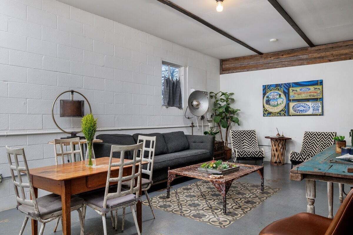 Ein Zimmer mit weißer Ziegelwand, einem grauen Sofa, einem Holzesstisch mit Stühlen, einem gemusterten Teppich und einer Decke mit sichtbaren Balken.