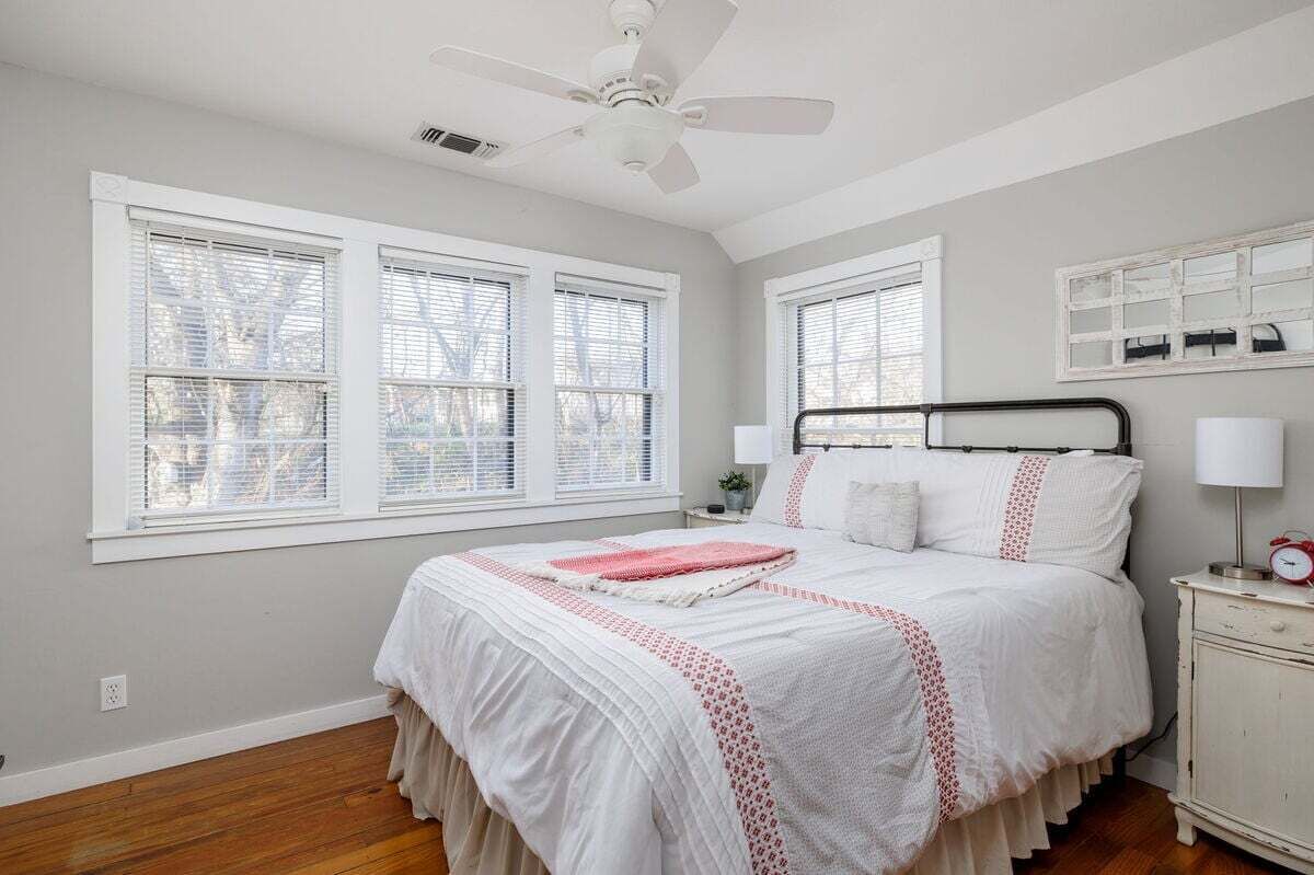A bedroom with a bed, a nightstand, a lamp, a ceiling fan, and three windows with blinds.