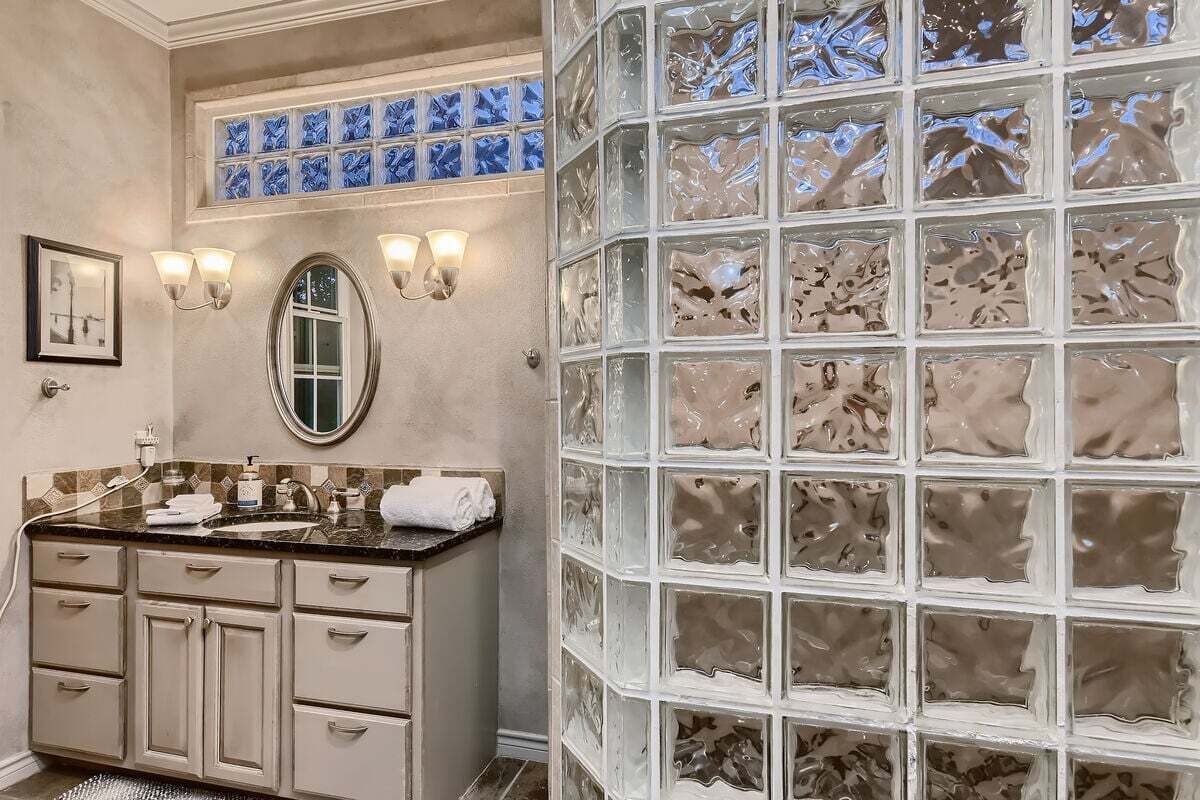 A bathroom with a vanity, a mirror, wall sconces, and a decorative glass partition.