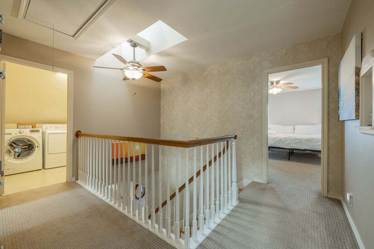Un cuarto de lavado con lavadora y secadora, una escalera con barandilla blanca, y un dormitorio con cama y ventilador de techo.