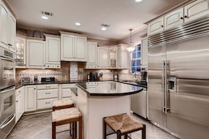 A kitchen with white cabinets, a stainless steel refrigerator, and a central island with stools. - Hill Country Retreat Private Pool Hot Tub Sleeps to 12 (Dripping Springs)
