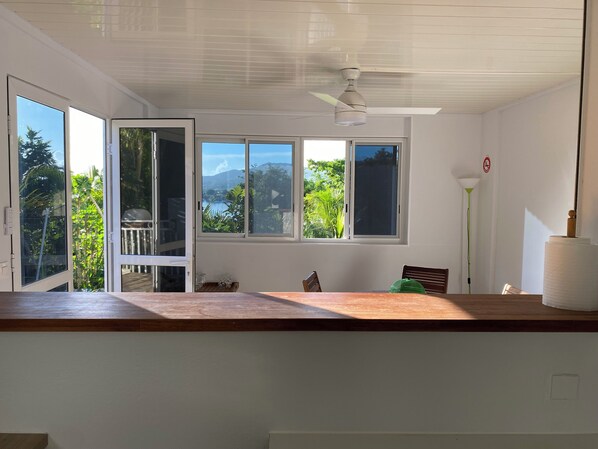 Ein Zimmer mit Holztisch, Deckenventilator und großen Fenstern mit Blick auf Grünflächen und Berge.