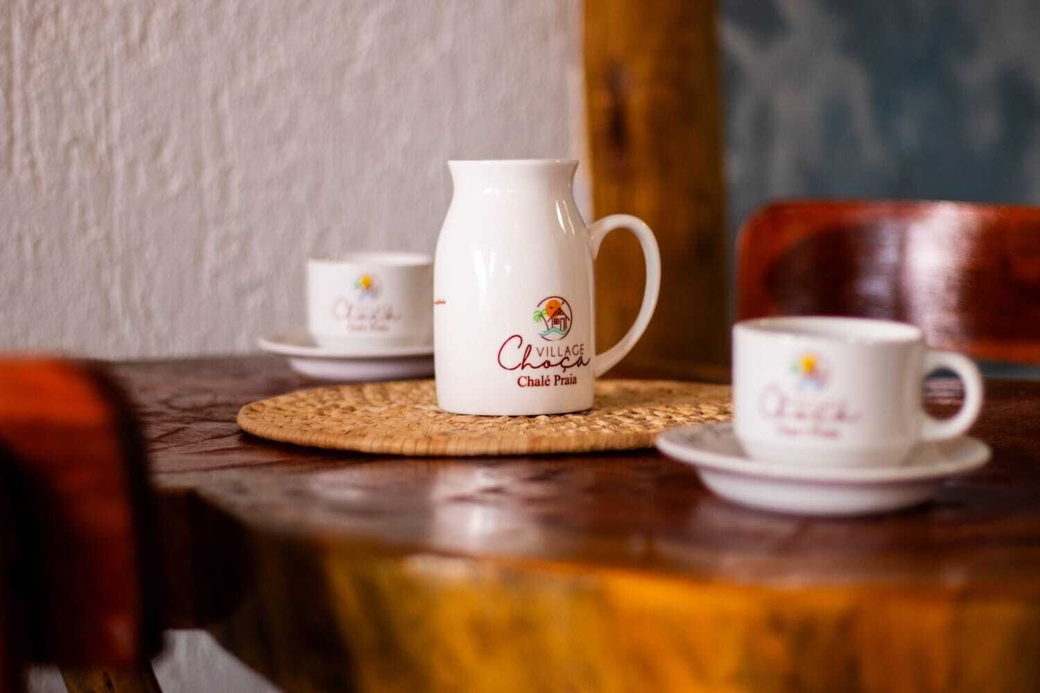 Una mesa con una jarra blanca y dos tazas blancas con logotipos, sobre un mantel tejido.