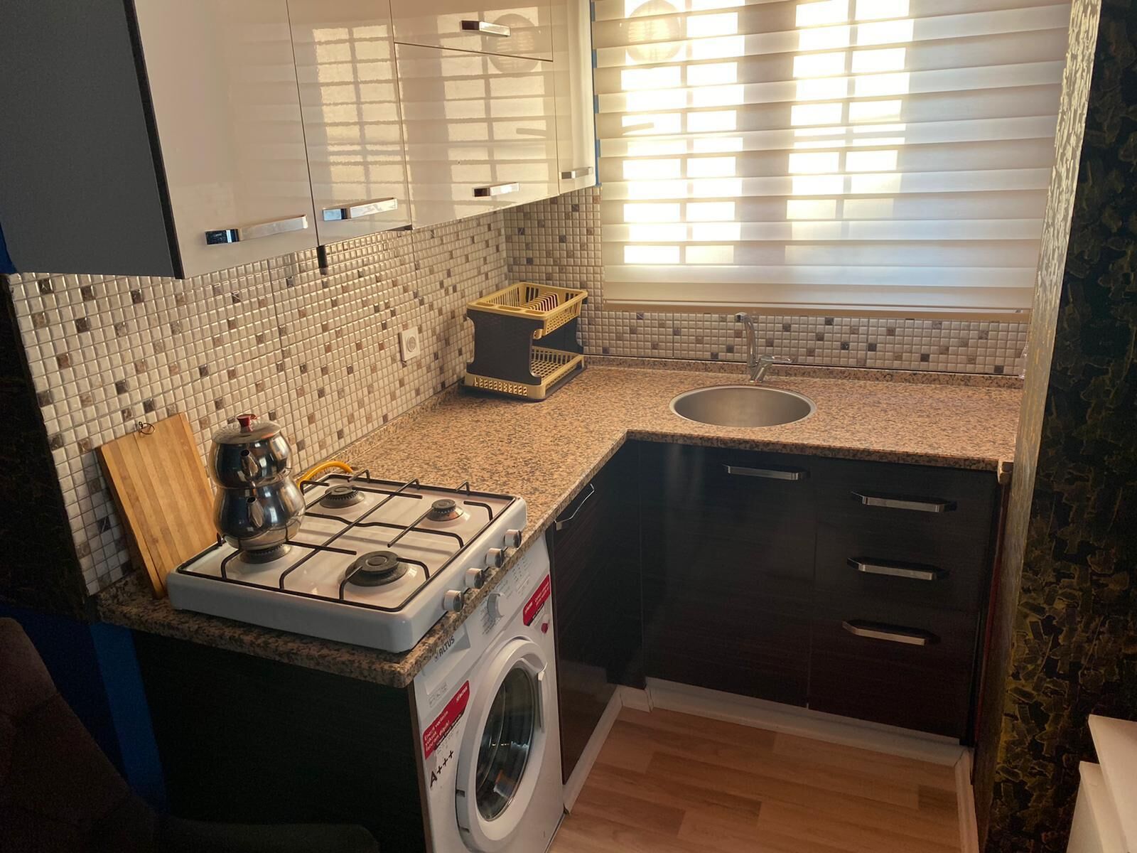 A compact kitchen with a gas stove, a built-in washing machine, a sink, and a window with blinds.