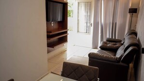 A living room with a sofa, a TV mounted on a wooden cabinet, a dining table, and a view of the outdoors through a door. - Casa Confortavel e Completa em Bariri (Bariri)