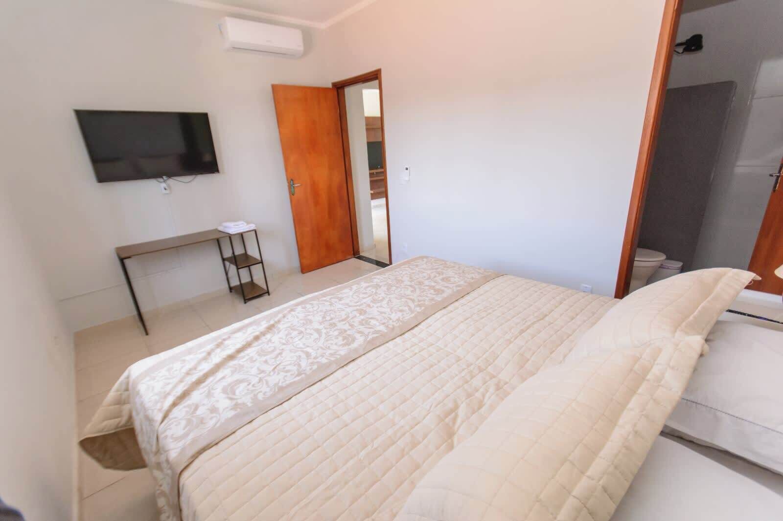 A bedroom with a bed, a TV mounted on the wall, a wooden door, and a small table with a shelf.