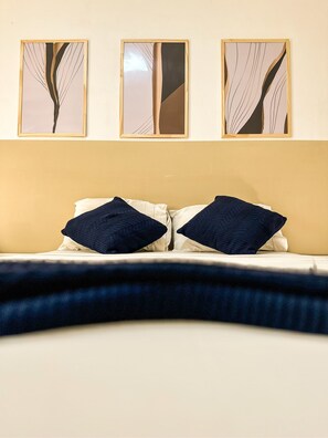 Een bed met twee donkere kussens, een lichtkleurig hoofdbord en drie ingelijste kunstwerken erboven.