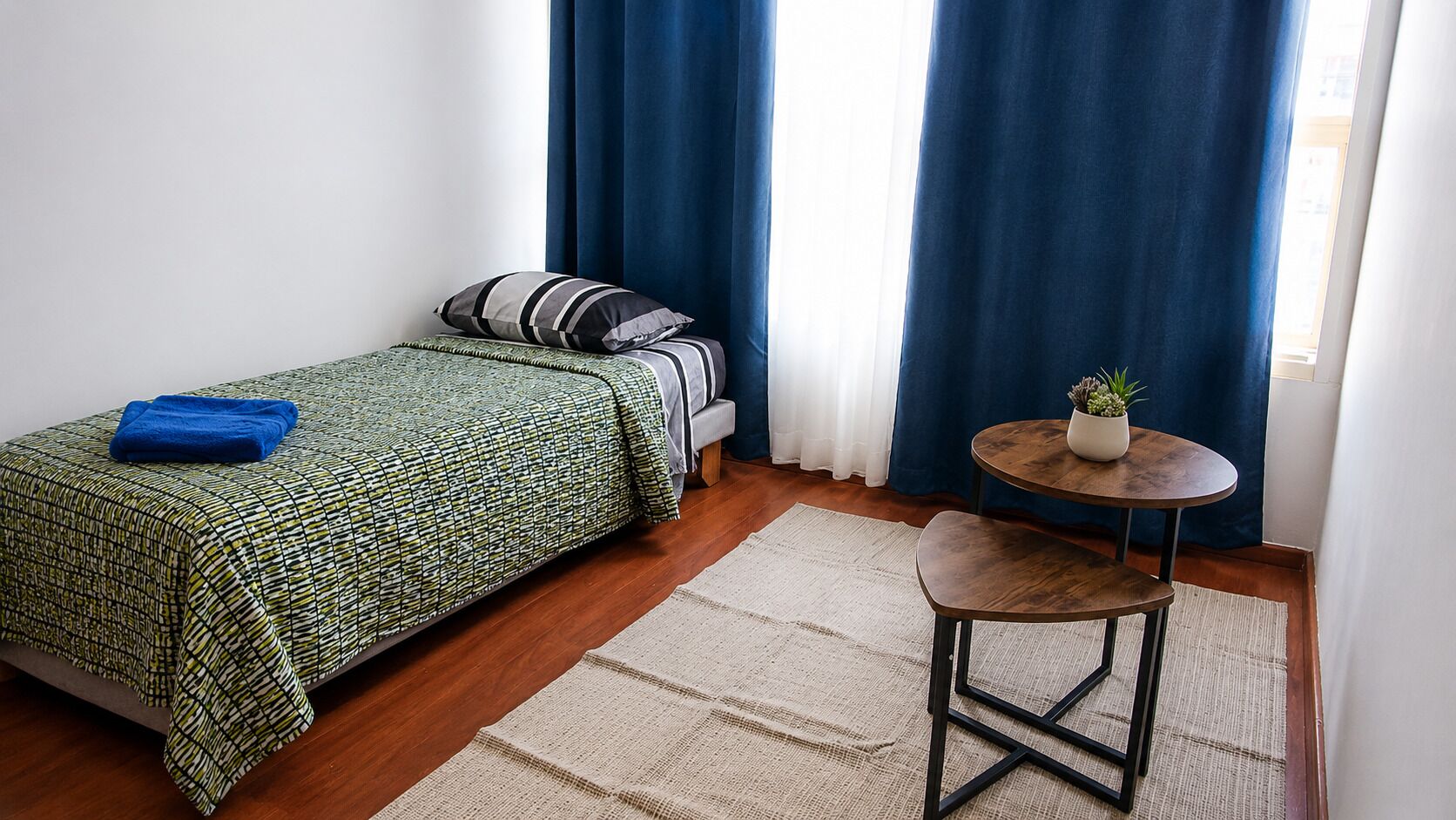 Un dormitorio con cama, una mesa redonda con una planta y una cortina azul.