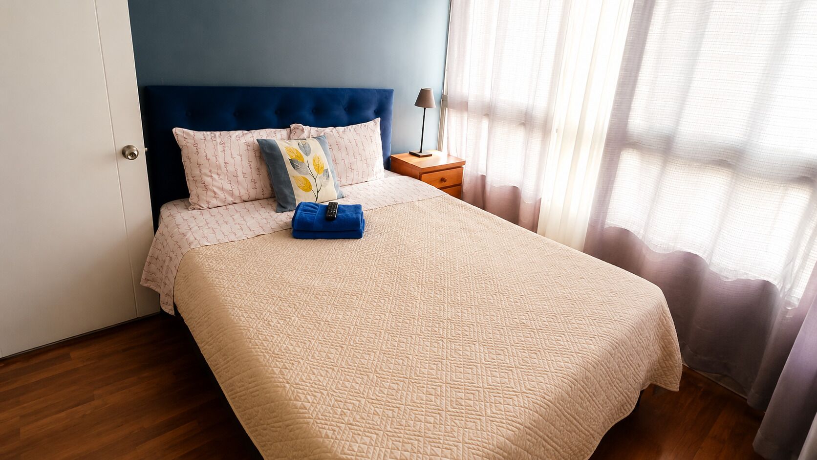 Ein ordentlich bezogenes Bett mit blauem Kopfteil, ein Nachttisch mit Lampe und ein Fenster mit durchsichtigen Vorhängen.