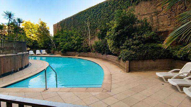 A swimming pool with a curved edge, surrounded by a tiled deck. There are lounge chairs and a brick wall with greenery.