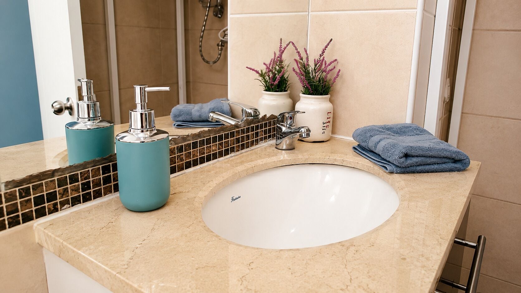 Un baño con un lavamanos blanco, dos grifos cromados, una toalla azul y dos jarrones blancos con flores rosadas.