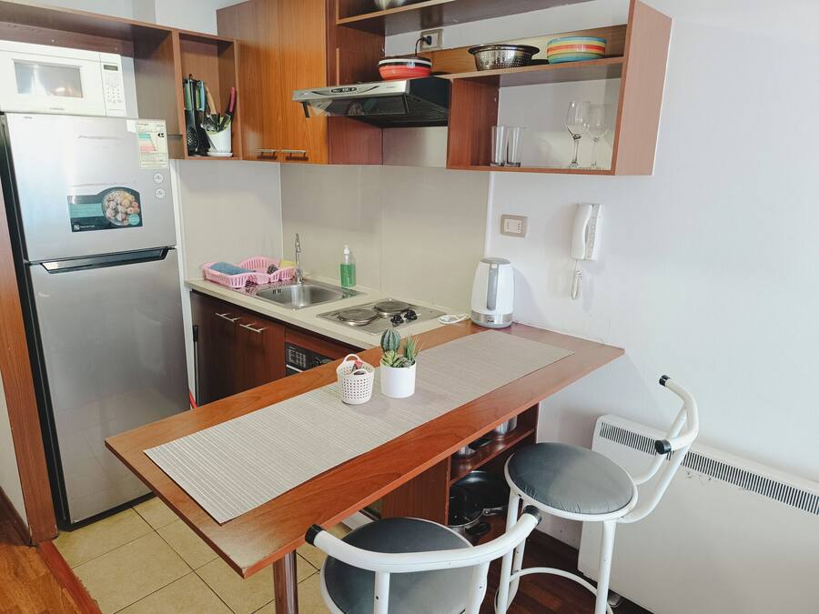 Cozinha compacta com geladeira, micro-ondas, pia e um pequeno espaço de jantar com mesa e duas cadeiras.