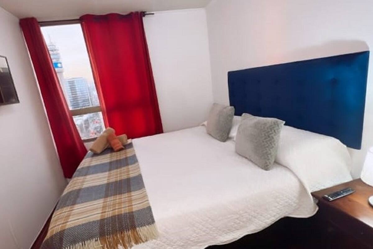 Una habitación de hotel con cama, almohadas, mesita de noche, control remoto y televisor montado en la pared.