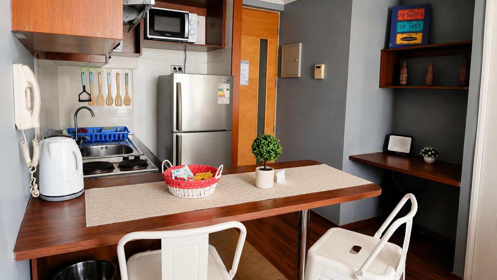 Cocina compacta equipada con microondas, refrigerador, hervidor y un pequeño comedor con sillas y una mesa.
