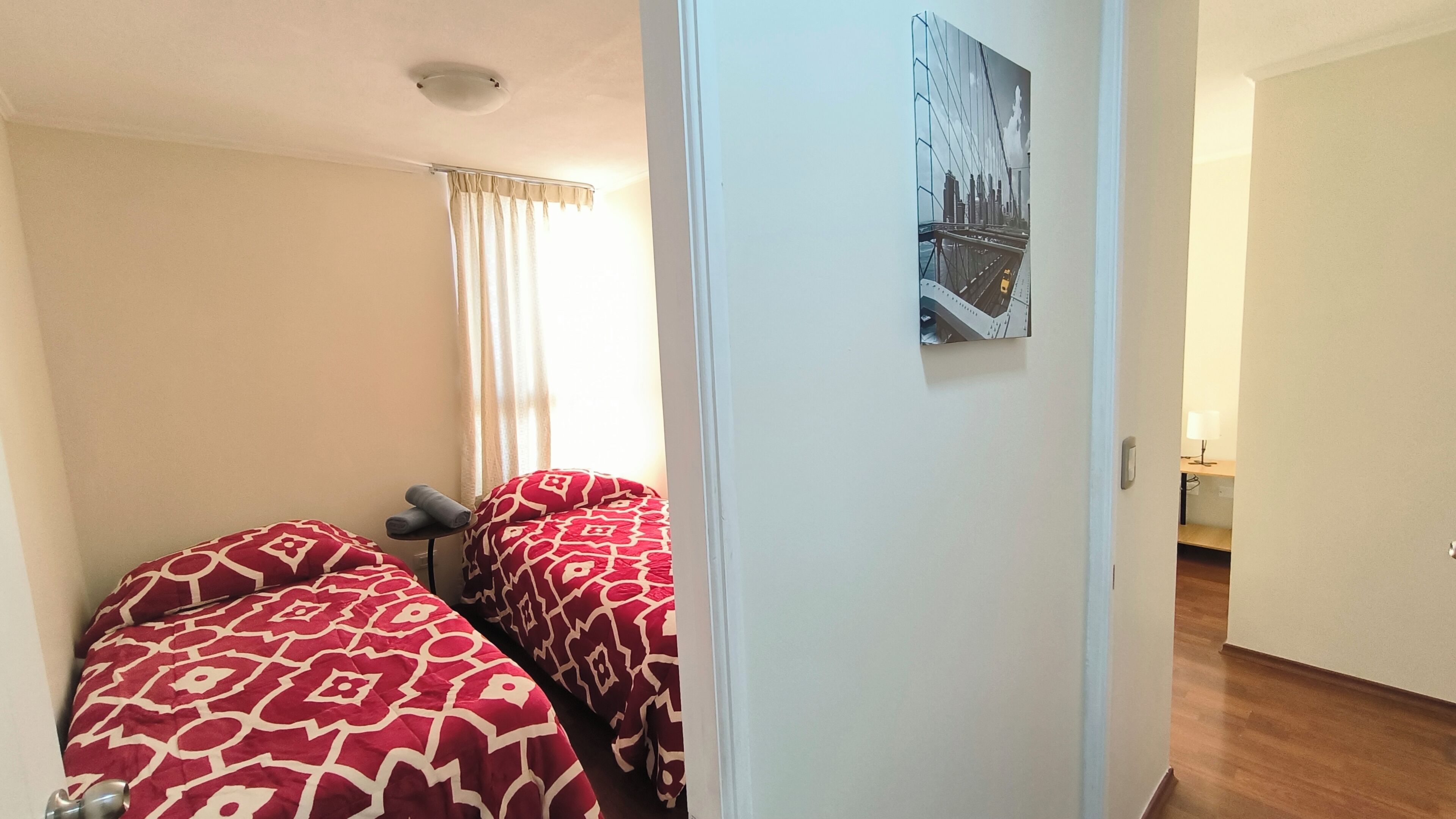 A room with two beds, a window with curtains, a door, and a cityscape painting on the wall.