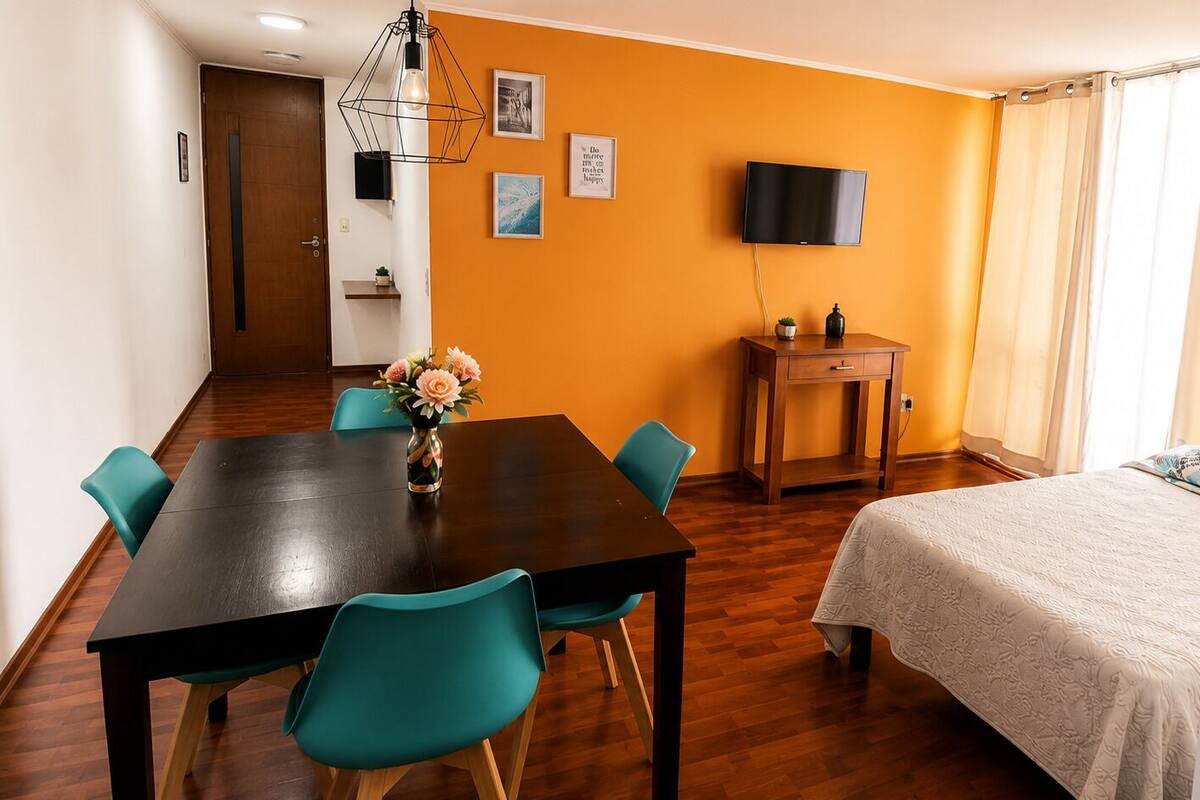 Habitación de hotel con una mesa de comedor de madera, sillas color turquesa, una cama con colcha blanca, un televisor en la pared y una pequeña mesita de madera con una planta.