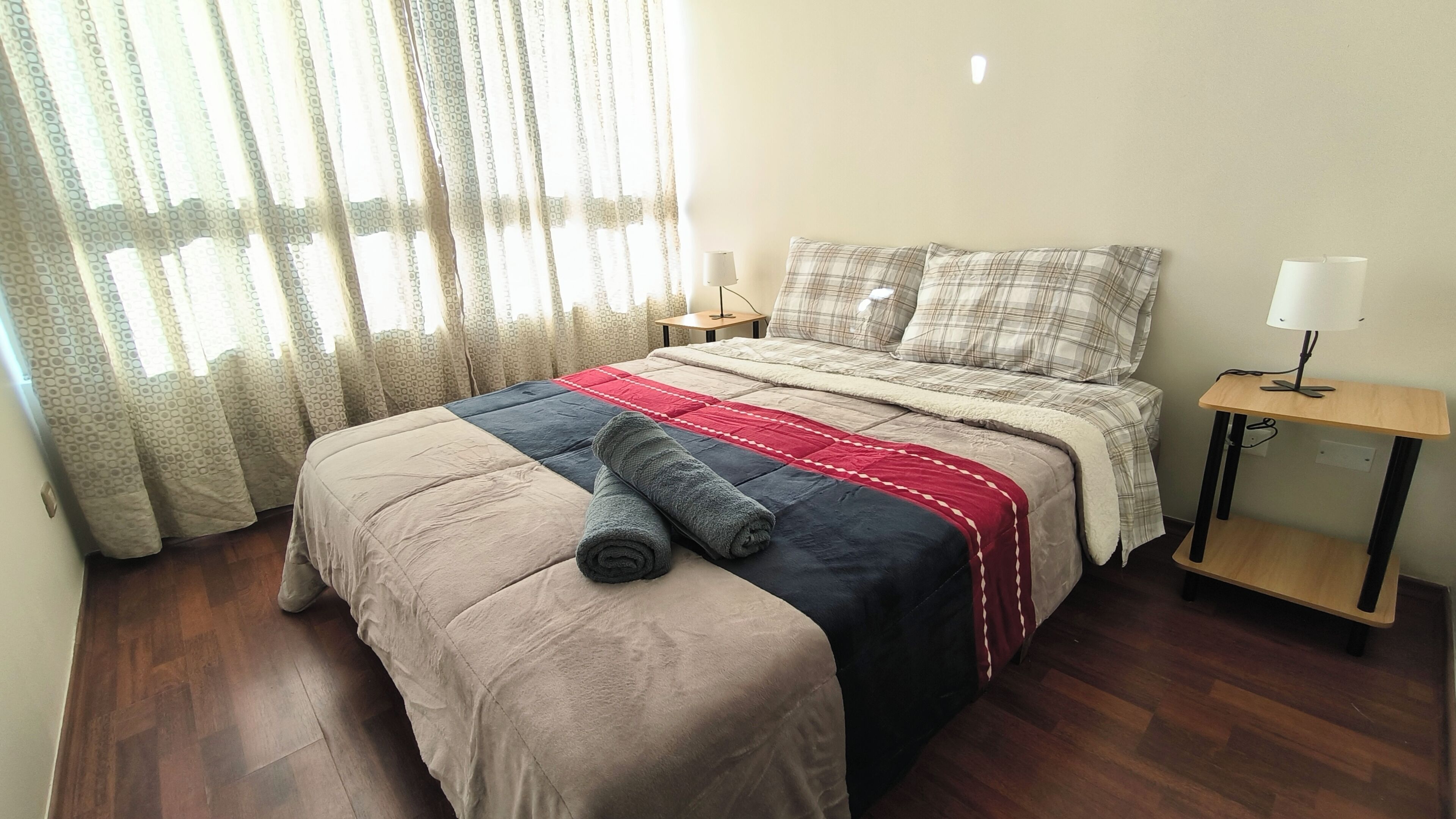 Una cama bien hecha con un edredón rojo y azul, una toalla enrollada y una mesita de noche de madera con una lámpara.