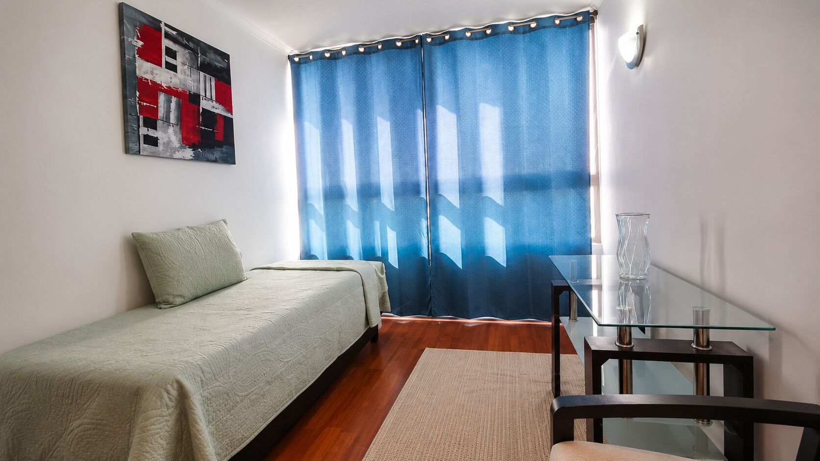 Una cama individual con una colcha verde, una mesita con superficie de vidrio y una cortina azul.