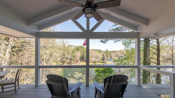 A covered porch with two Adirondack chairs, a table, and a view of a lake surrounded by trees. - LAKEFRONT PROPERTY l DANCING BEAR (Lake Toxaway)