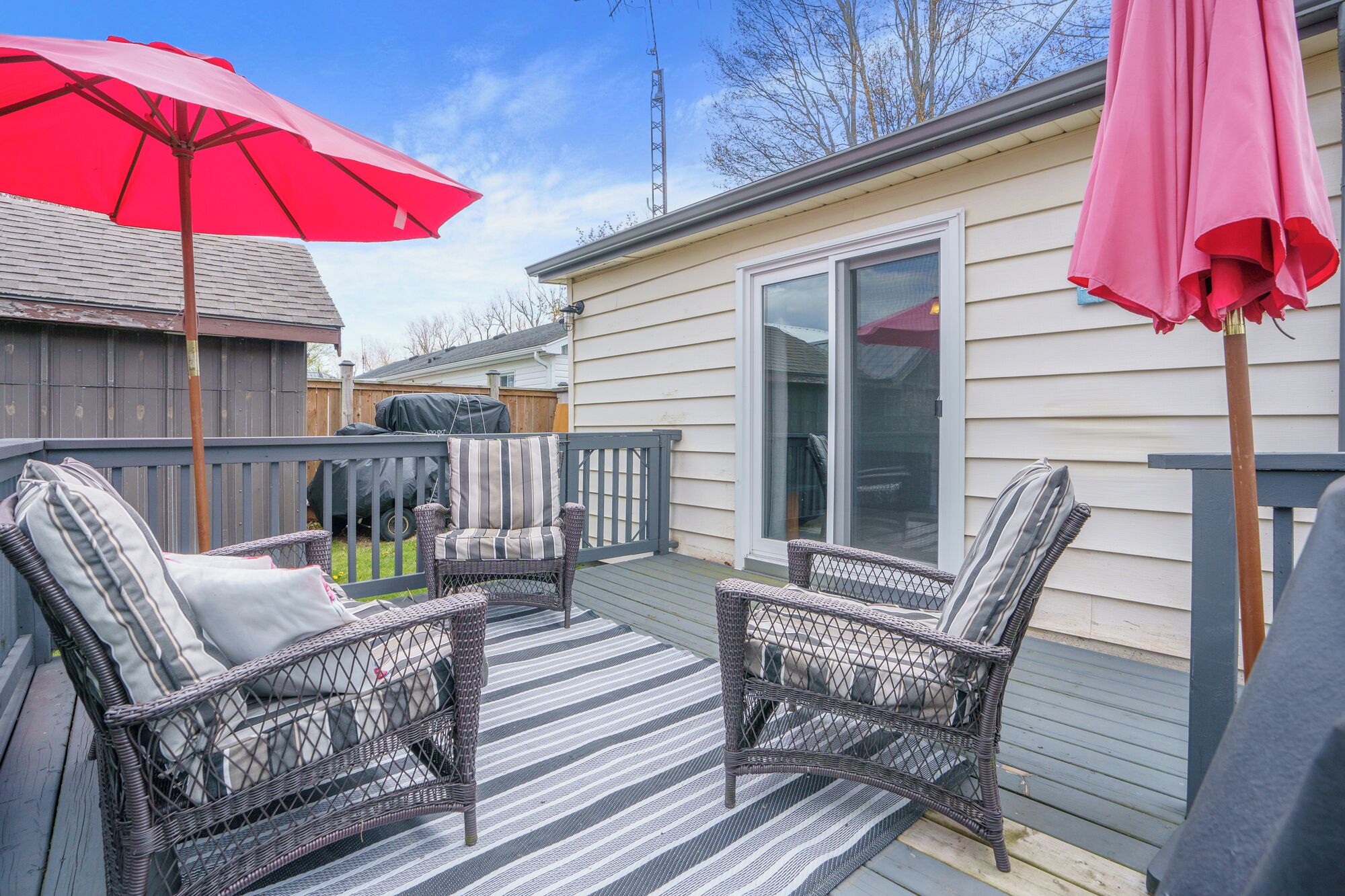 Une terrasse avec des fauteuils en osier et un tapis à rayures, un barbecue abrité et une porte-fenêtre coulissante.