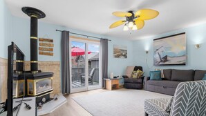 Un salón con chimenea, un sofá, un sillón estampado, un ventilador de techo y vistas a través de un ventanal.