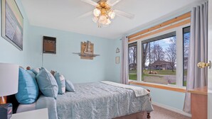Un dormitorio con cama, ventilador de techo, ventana con vistas al exterior y repisa en la pared.