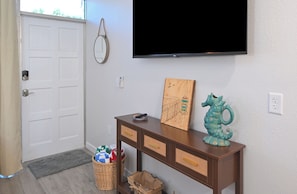 Ein Zimmer mit einer weißen Tür, einem flachbildfernseher an der Wand, einem Holzkonsolentisch mit Schubladen, einem Korbwäschekorb und einer kleinen Mülltonne.