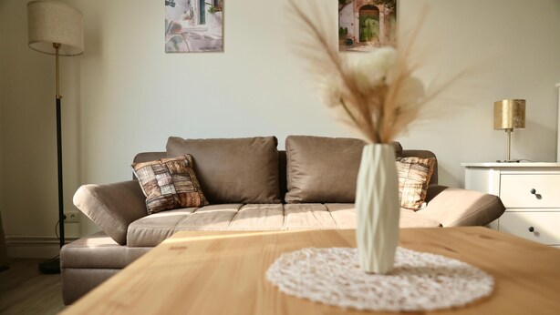 Een woonkamer met een bank, een houten salontafel, een vaas met gedroogde planten en een lamp.