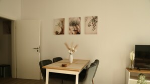 Een eetruimte met een houten tafel, zes stoelen, een vaas met gedroogde bloemen en drie kunstwerken in lijsten aan de muur.