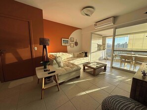 A living room with a sofa, a coffee table, a lamp, and a sliding glass door leading to a balcony. - @cyclinn.natureza | All Time 88 T2 (100m praia) (Riviera de São Lourenço)
