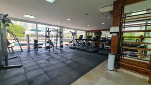 A gym with various exercise equipment, large windows, and a view of buildings and greenery outside. - @cyclinn.natureza | All Time 88 T2 (100m praia) (Riviera de São Lourenço)