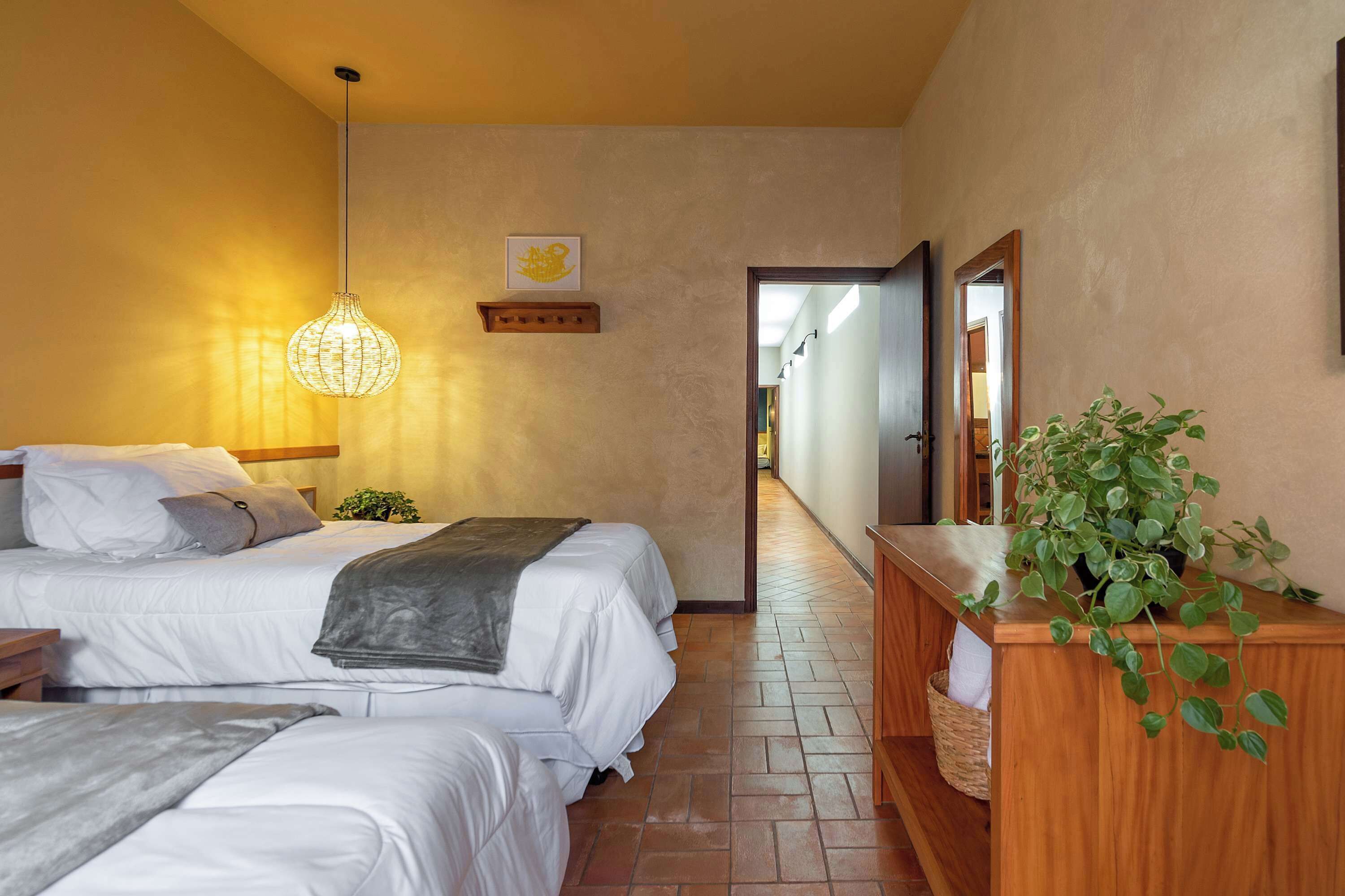 Habitación de hotel con dos camas, una mesita de noche, una lámpara colgante, una planta pequeña y un aparador de madera con espejo.