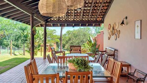 An outdoor dining area with a glass table, wooden chairs, woven pendant lights, and a view of the garden. - Rancho da Vó Vera | Recém Reformado e Bem Equipado (Bariri)
