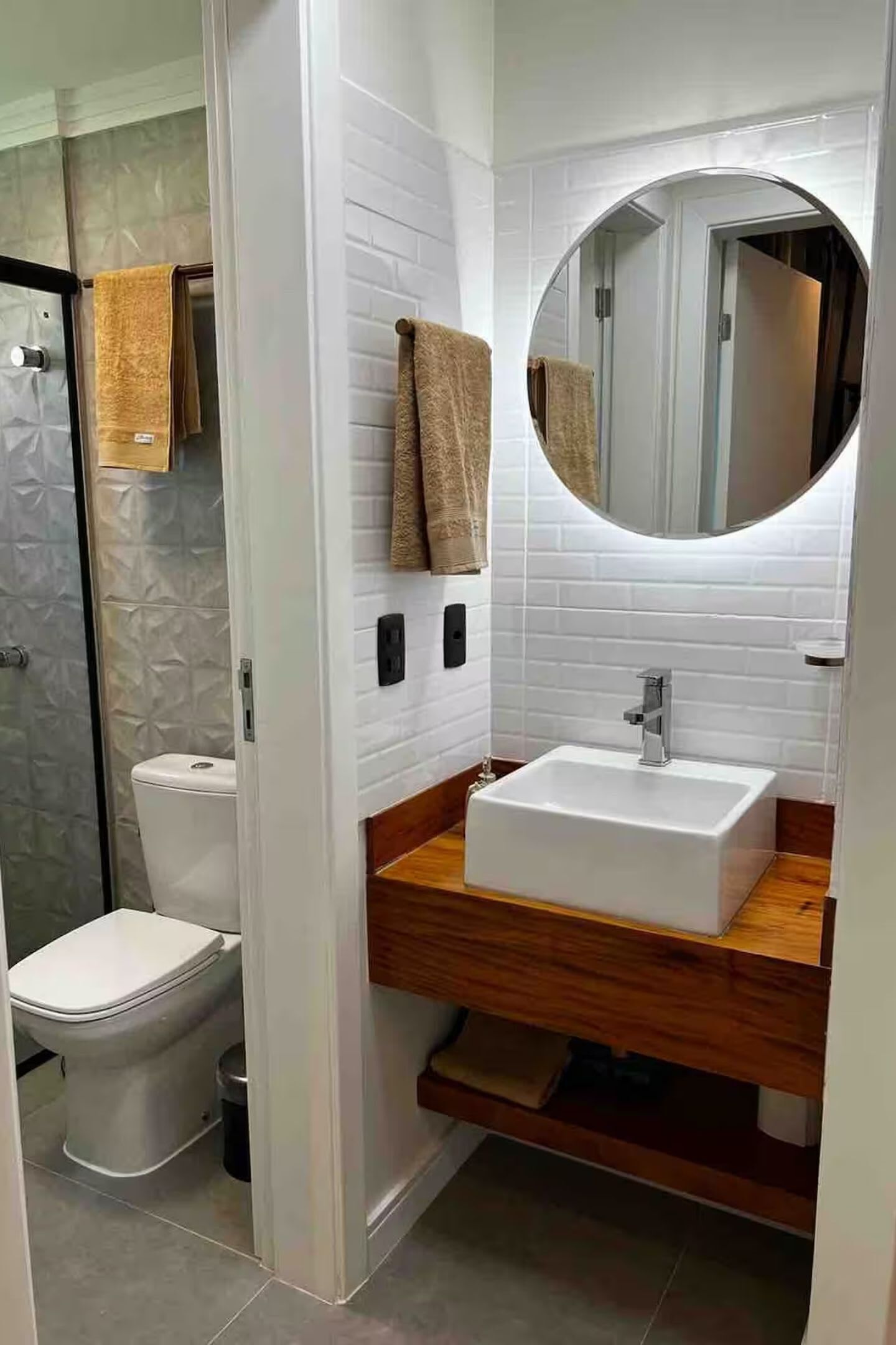 Une salle de bain avec des toilettes blanches, un meuble-lavabo en bois, un miroir rond et un porte-serviettes.