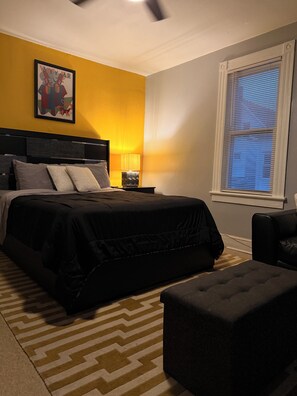 Ein Schlafzimmer mit einem Bett, Nachttischen, einer Lampe, einem Fenster mit Jalousien und einem gemusterten Teppich.