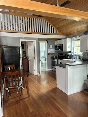 A kitchen with wooden flooring, a dining area with a table and chairs, a refrigerator, and a ceiling fan.