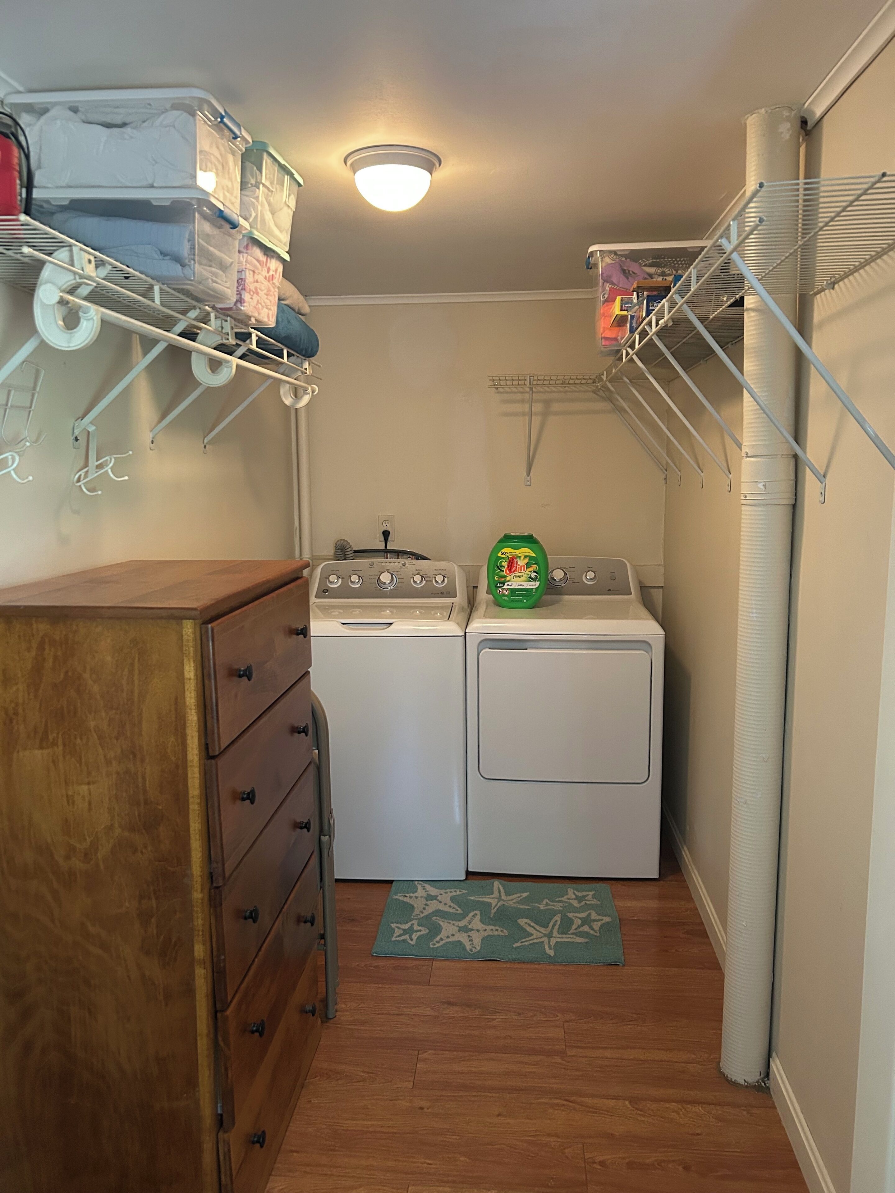 Une buanderie équipée d’un lave-linge et d’un sèche-linge, d’une commode en bois, d’étagères en métal et d’un luminaire au plafond.
