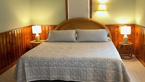 A bedroom with a large bed, wooden paneling, two bedside lamps, and a window with a closed shutter. - Pines Cottage – Yarmouth w/ Private Beach Rights (Yarmouth)