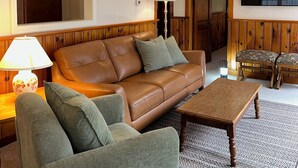 A living room with a brown leather sofa, a green armchair, a wooden coffee table, a lamp, and a television mounted on the wall. - Pines Cottage – Yarmouth w/ Private Beach Rights (Yarmouth)