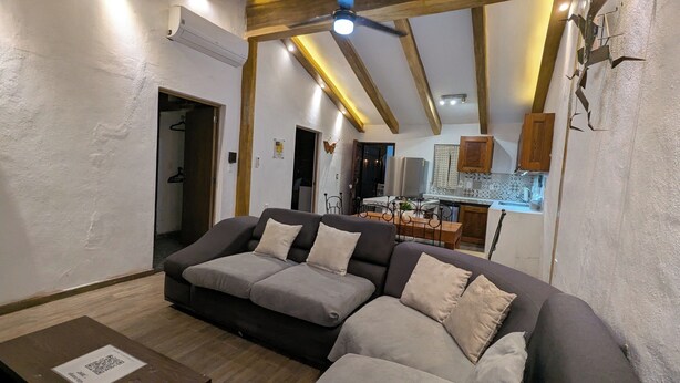 Un salon avec un canapé, des poutres en bois, un ventilateur de plafond et, au fond, une cuisine.