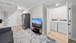 Een moderne woonkamer met een flatscreen televisie, een grijze bank, een vloerkleed met patroon, een keukenhoek met een koelkast, en een wasruimte met een gestapelde wasmachine en droger.