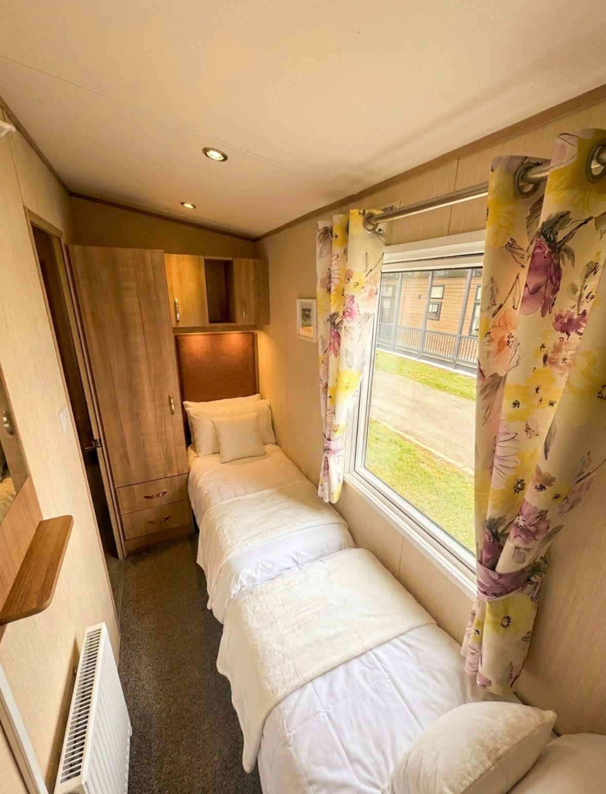 A narrow room with two beds, wooden cabinets, a radiator, and a window with floral curtains.