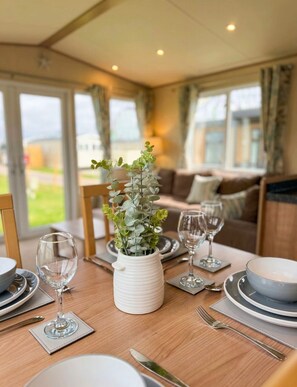 A dining area with a table set for four, a white vase with greenery, and a sofa in the background. - Fantasy Island Resort, The Sunnydale C6 Eastgate - Coastal Caravan Breaks (Ingoldmells)