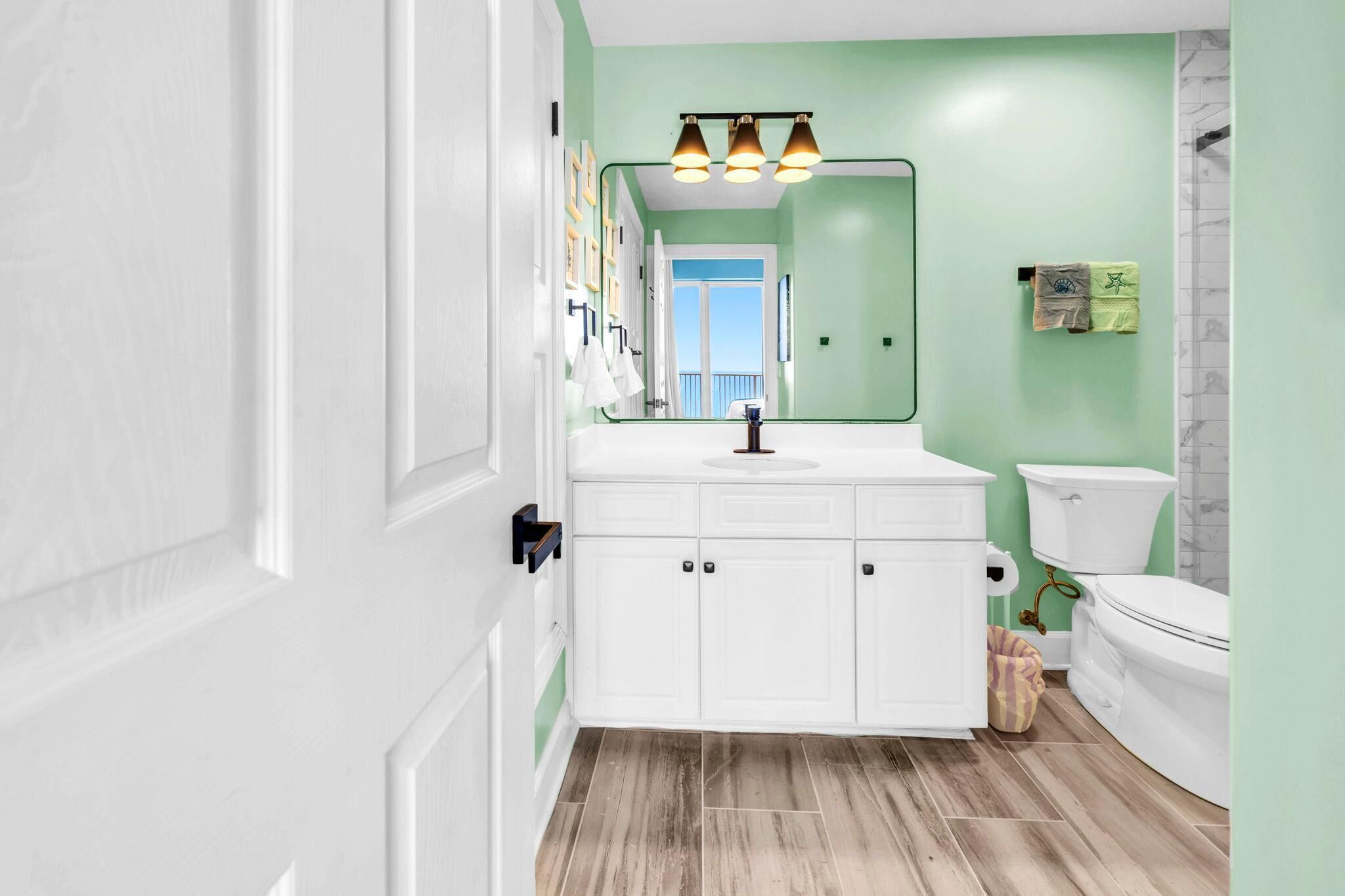 Un baño con armarios blancos, pared verde, inodoro blanco y espejo con tres luces.