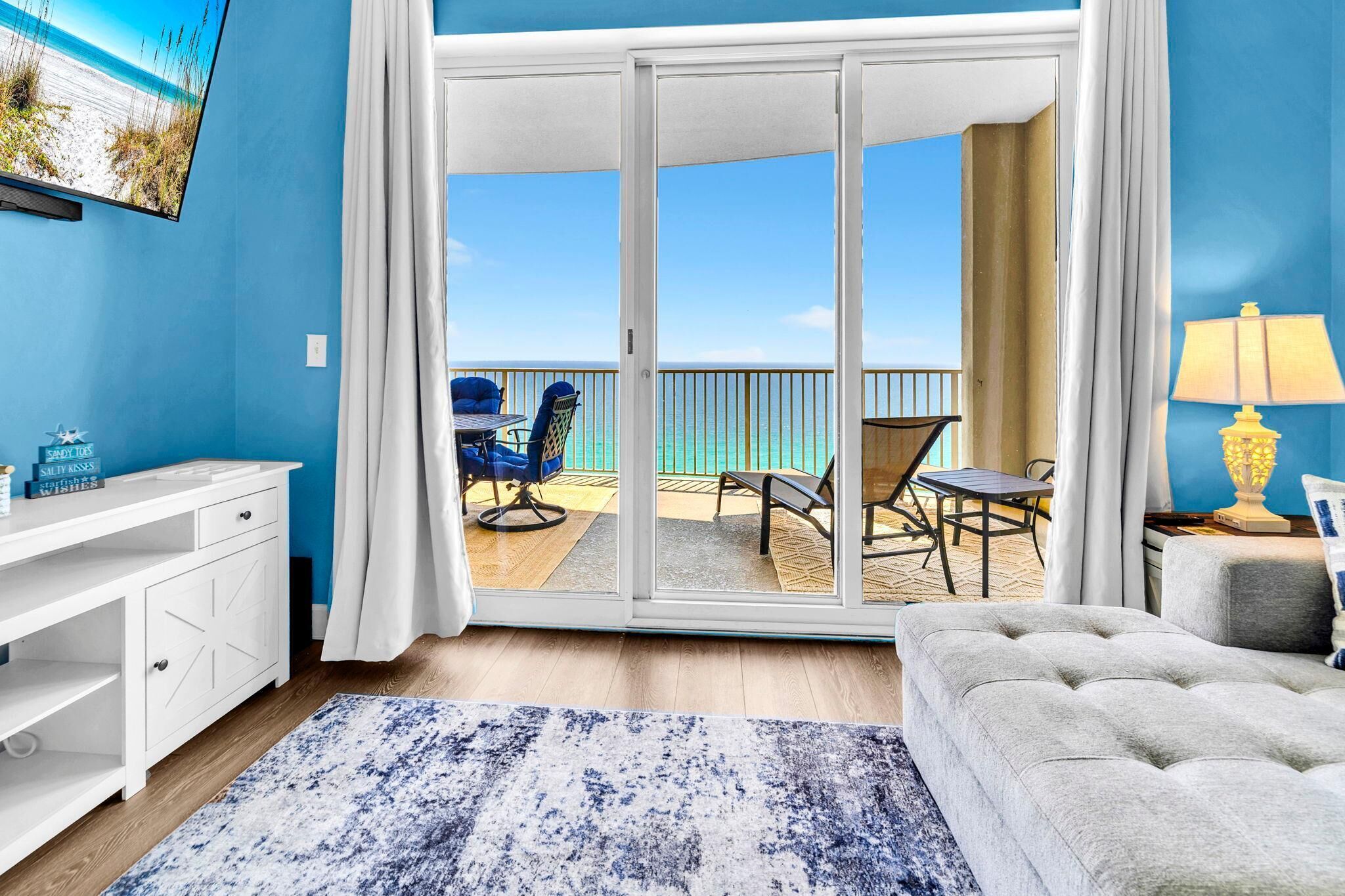 Habitación con puerta de vidrio corredera que da a un balcón con vistas a la playa, un escritorio blanco, un sofá gris y una lámpara.