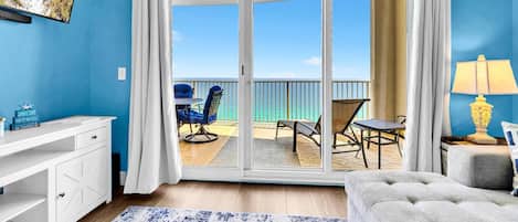 Habitación con puerta de vidrio corredera que da a un balcón con vistas a la playa, un escritorio blanco, un sofá gris y una lámpara.