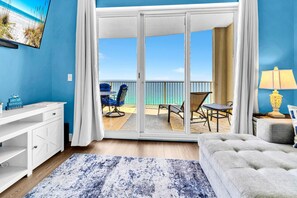 Ein Zimmer mit einer Schiebeglastür, die auf einen Balkon mit Blick auf den Strand führt, einem weißen Schreibtisch, einem grauen Sofa und einer Lampe.
