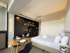 Moderni hotellihuone, jossa on sänky, ruokapöytä tuoleineen, keittiö sekä ilmastointilaite.