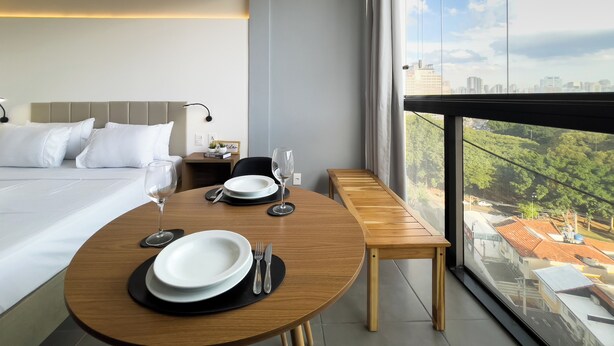 A modern hotel room with a large bed, a dining table set with plates and cutlery, a bench, and a city view through large windows. - VN Alvorada (São Paulo)